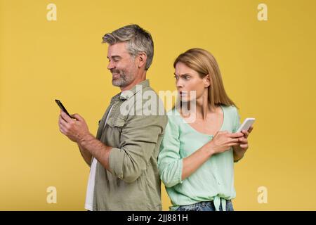 Schockierte Frau, die ihren glücklichen Mann mit dem Smartphone ausspioniert und sich über gelben Hintergrund zurück an den Rücken stellt Stockfoto