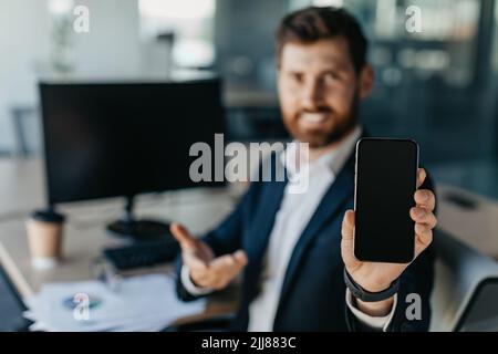 Mobiles Angebot. Geschäftsmann zeigt leeres Smartphone, während er am Schreibtisch im Büroinneren sitzt, selektive Fokussierung, Mockup Stockfoto