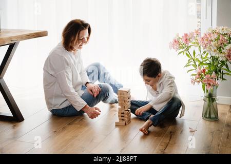 Mutter mit Kind Sohn sitzen auf dem Boden spielen ein Brettspiel der körperlichen Geschicklichkeit Turm. Zerstörte den Turm. Über sonniges Fenster. Jenga, Janga von Stockfoto
