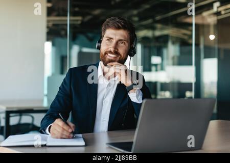 Ein glücklicher, nachdenklicher Geschäftsmann mit Headset, der am Laptop und im Notebook arbeitet und am Schreibtisch im Büroinneren sitzt Stockfoto