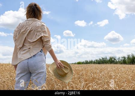 Frau, die im Sommer auf einem Weizenfeld posiert. Frau, die einen Hut in der Hand vor dem Hintergrund eines goldenen Weizenfeldes hält. Der Blick von hinten. Stockfoto