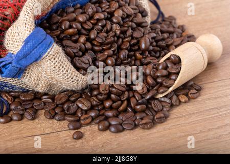 Geröstete schwarze Kaffeebohnen mit einem Holzlöffel, die aus dem Sack auf den Tisch gelangen. Foto für ein Café, Café oder Restaurant. Stockfoto