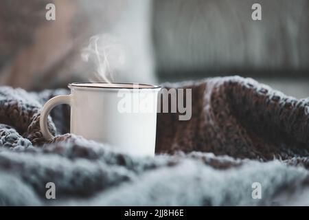 Weiße emaillierte Tasse heißen, dampfenden Kaffees auf einem gemütlichen Bett, umgeben von Kissen und Strickdecken. Selektiver Fokus mit unscharfem Hintergrund. Stockfoto