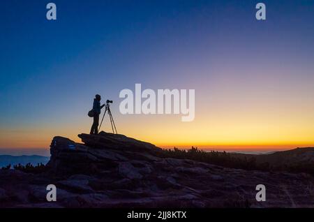 Silhouette des Fotografen bei Sonnenuntergang. Der Fotograf mit dem Stativ fotografiert hoch in den Bergen Stockfoto