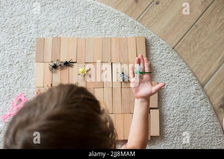 Kaukasisches Mädchen, 5 Jahre alt, spielt mit Holzblöcken und Spielzeugtieren auf dem Boden und sitzt auf beigefarbenem Teppich. Draufsicht, selektiver Fokus. Stockfoto