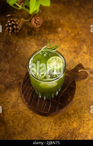 Frisch aus Eis im Glas mit Zitronenscheibe und Rosmarin zubereiteter Gemüsesaft, auf rustikalem Holztablett und authentischem Hintergrund mit Minipflanzen in CO Stockfoto