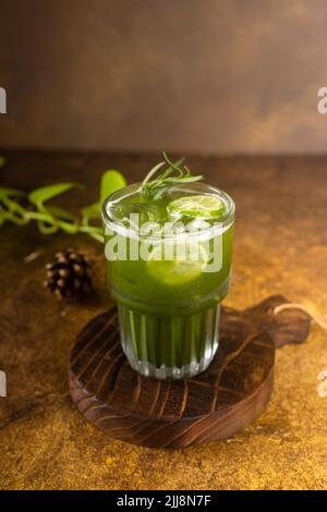 Frisch aus Eis im Glas mit Zitronenscheibe und Rosmarin zubereiteter Gemüsesaft, auf rustikalem Holztablett und authentischem Hintergrund mit Minipflanzen in CO Stockfoto