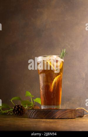 Frischer Zitronentee aus Eis im Glas mit Zitronenscheibe und Rosmarin, auf rustikalem Holztablett und authentischem Hintergrund Stockfoto
