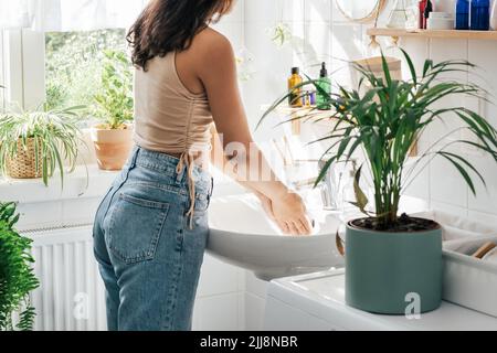 Anonyme junge hispanische Frau, die sich im weißen Badezimmer mit grünen Pflanzen die Hände wascht. Wellness-Konzept Stockfoto