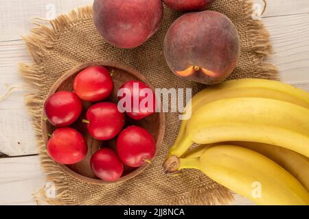 Ein paar süße, saftige Kirschpflaumen, zwei Pfirsiche und ein paar Bananen mit Jute-Serviette auf einem Holztisch, Nahaufnahme, Draufsicht. Stockfoto