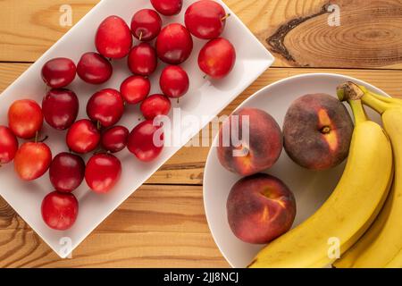 Mehrere süße, saftige Kirschpflaumen, drei Pfirsiche und ein Bund Bananen mit weißen Keramikutensilien auf einem Holztisch, Nahaufnahme, Draufsicht. Stockfoto