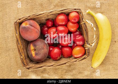 Ein paar süße saftige Kirschpflaumen, eine Banane und zwei Pfirsiche in einem Weidenkorb auf Sackleinen, Nahaufnahme, Draufsicht. Stockfoto