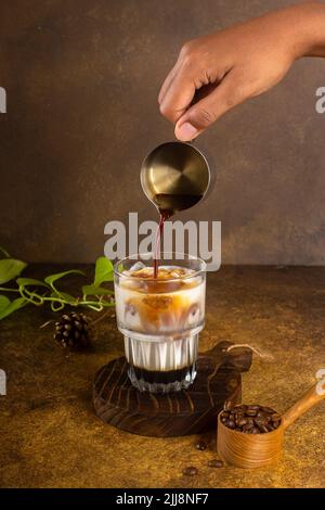 eiskaffee mit Kondensmilch auf rustikalem Holzuntersetzer und mit grünen Pflanzen, volle Kaffeebohne auf Vintage-Holzlöffel auf altem rustikalem Hintergrund Stockfoto