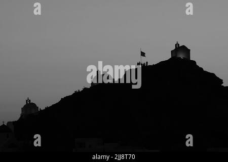 Touristen erkunden die malerischen kleinen Kapellen auf der atemberaubenden Insel iOS Greece, während die Sonne schwarz-weiß untergeht Stockfoto