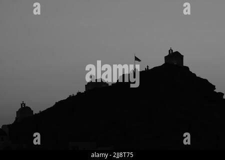 Touristen erkunden die malerischen kleinen Kapellen auf der atemberaubenden Insel iOS Greece, während die Sonne schwarz-weiß untergeht Stockfoto