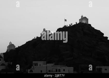 Touristen erkunden die malerischen kleinen Kapellen auf der atemberaubenden Insel iOS Greece, während die Sonne schwarz-weiß untergeht Stockfoto