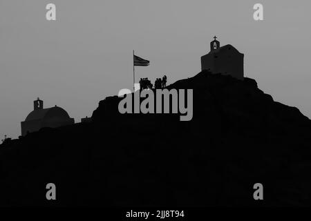 Touristen erkunden die malerischen kleinen Kapellen auf der atemberaubenden Insel iOS Greece, während die Sonne schwarz-weiß untergeht Stockfoto