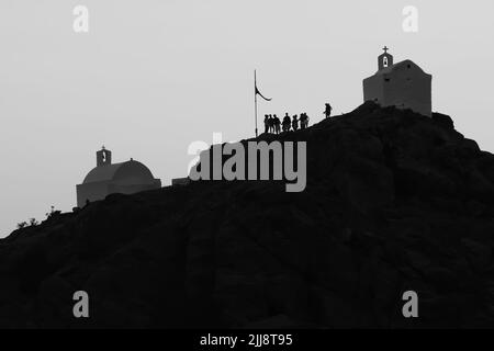 Touristen erkunden die malerischen kleinen Kapellen auf der atemberaubenden Insel iOS Greece, während die Sonne schwarz-weiß untergeht Stockfoto