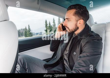 Kaukasischer junger erfolgreicher Mann, der ernsthaft auf dem Smartphone spricht, während er auf dem Rücksitz eines fahrenden Autos sitzt Stockfoto