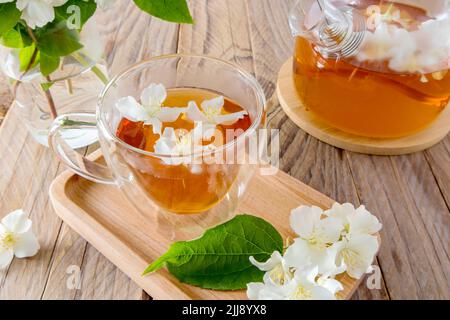 Eine Tasse natürlichen Jasmintee in einer Glastasse auf einem Holztablett mit weißen Blumen. Ein rustikaler Tisch mit einer Teekane und Jasminzweigen in einer Vase Stockfoto