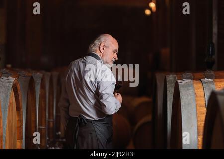 Kaukasischer Senior-Graubart-Winzer, der Rotwein verkostet, um seine Qualität und seinen Geschmack zu ermitteln, steht in der Nähe eines großen Eichenfasses Stockfoto