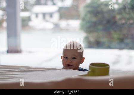 Neugierig, verspielt und schelmisch Kleinkind mit einem Finger in der Halterung beobachtet Eltern Bett interessiert an einer Kaffeetasse, blonde Junge in der hellen Stockfoto