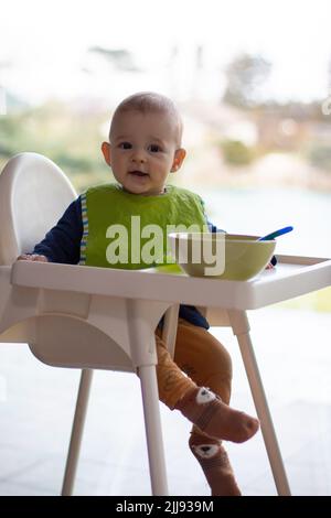 Kleines Kleinkind, das auf einem Hochstuhl isst, glückliches Baby, ein Vollkörperbild, lernt zu essen und hat Nahrung im Gesicht Stockfoto