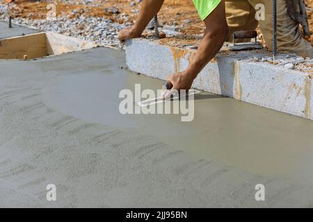 Es gibt einen Mauerarbeiter, der eine Stahlkelle hält, die Beton auf einen Zementboden glättet Stockfoto