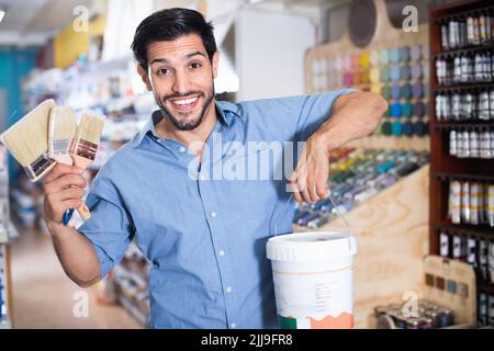 Mann, der mit Bürsten und Farbe zwischen den Regalen im Lacklager steht Stockfoto