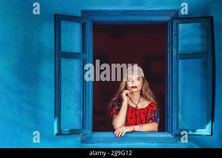 Porträt einer schönen Zigeunerin in traditionellem roten Kleid, die am Fenster steht – Glück und Horoskop-Hexenerzähler Stockfoto