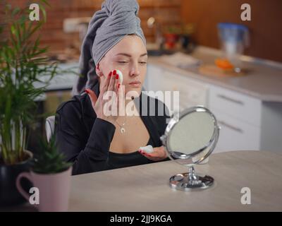 Genießen Sie makellose Haut . Friedliche Dame, die während der Eingriffe in der Küche mit dem Zustand ihrer Haut zufrieden war. Schöne kaukasische Frau putzt Gesicht mit reinigenden Schaum in Spiegel suchen. Stockfoto
