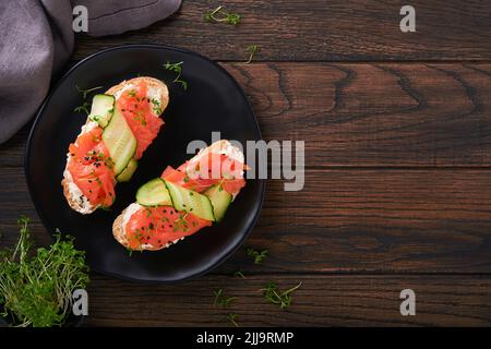 Sandwiches. Lachs-Toast mit Frischkäse, Gurke, schwarzem Sesam und Microgreens auf altem Holztisch-Hintergrund. Meeresfrüchte. Gesunde Ernährung. Fotografie Stockfoto