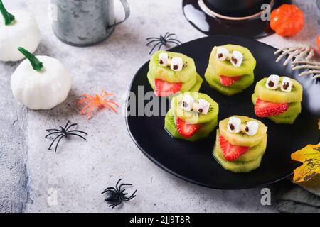 Gruselige grüne Kiwi-Monster für Halloween. Gesunde Früchte Halloween Leckereien. Halloween Party Kiwi, Erdbeere, Apfel und Sumpfmonster auf grauem ston Stockfoto