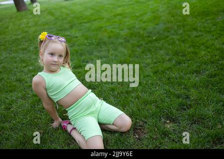 Ein kleines Mädchen im grünen Anzug sitzt auf dem Rasen im Park und posiert für die Kamera Stockfoto