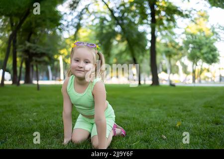 Ein kleines Mädchen im grünen Anzug sitzt auf dem Rasen im Park und posiert für die Kamera Stockfoto