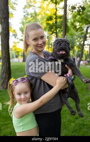 Mutter mit Tochter im Park Mutter hält eine französische Bulldogge in den Armen Stockfoto