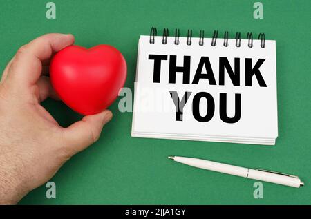 Auf der grünen Oberfläche ist ein Stift, ein Notizblock mit der Aufschrift - Danke. Ein Mann hat ein Herz in seinen Händen. Stockfoto