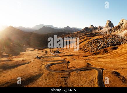 Malerische Luftaufnahme auf kurvenreicher Straße im herbstlichen Bergtal bei Sonnenuntergang. Das goldene Abendlicht beleuchtet die Berge und das orangefarbene Gras. Giau-Pass, Dolomiten, Dolomiten, Italien Stockfoto