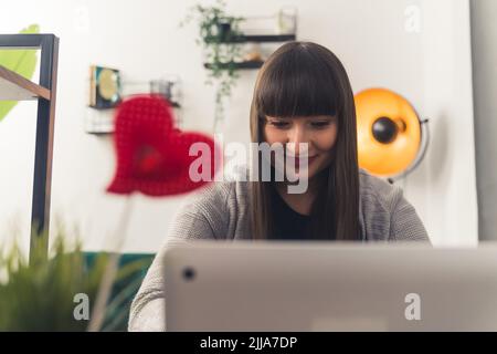Lächelnde weiße Frau mit dunklen Haaren und knallt in bequemen Kleidern, die auf einem Laptop im Wohnzimmer arbeiten. Innenaufnahme. Hochwertige Fotos Stockfoto
