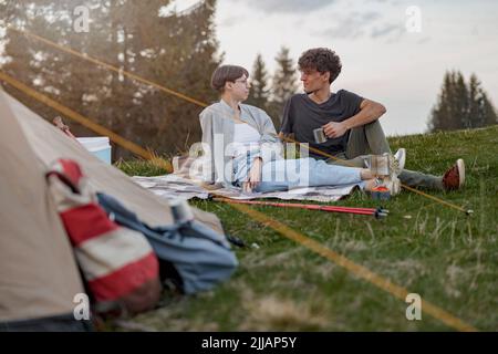 Junge kaukasische glückliche Mann und Frau sitzen auf Gras im Zelt. Bergtour. Stockfoto