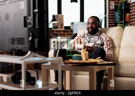 Junge Erwachsene essen Fast-Food-Delivery-Mahlzeit zu Hause, sehen Film im Fernsehen und genießen Freizeit. Es werden Burger vom Take-out-Paket, Chips und Bierflaschen im tv-Film serviert. Stockfoto