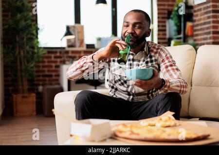 Fröhlicher Erwachsener, der alkoholisches Bier aus der Flasche trinkt und eine Schüssel mit Chips hält, während er sich im Fernsehprogramm einen Film ansieht. Genießen Sie Getränke mit Lieferservice zum Mitnehmen, Snacks und Unterhaltung. Stockfoto