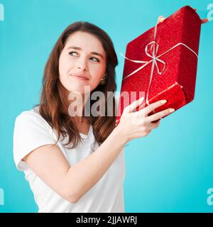 Ich Frage mich, was das sein könnte. Studioaufnahme einer attraktiven jungen Frau, die versucht, herauszufinden, was ihr Geschenk vor blauem Hintergrund ist. Stockfoto