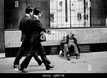 Zwei asiatische/chinesische Männer laufen im Schritt perfekt an einem verwelkenden Mann vorbei, der 1969 in einer Milchkiste vor dem Schneiderbüro in der Gerrard Street, Soho, London, hockt Stockfoto