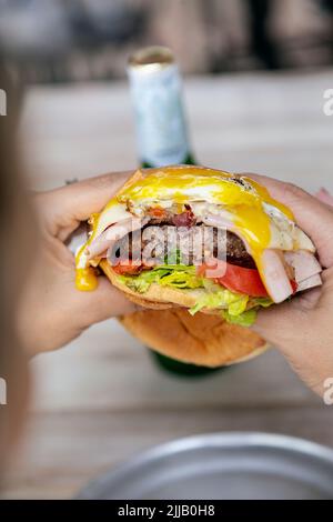 Cheeseburger mit Schinken, Spiegelei, Salat, Tomate und Zwiebel, gebissen von einer Person, die ihn in den Händen hält. Stockfoto