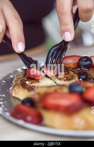Hände mit Plastikbesteck, um in einem Restaurant Blaubeer- und Erdbeerpfannkuchen zu schneiden Stockfoto