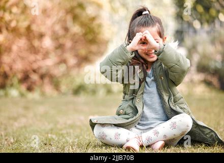 Ich liebe es draußen zu spielen. Ein kleines Mädchen formt sich eine Herzform über ihrem Auge, während es draußen sitzt. Stockfoto