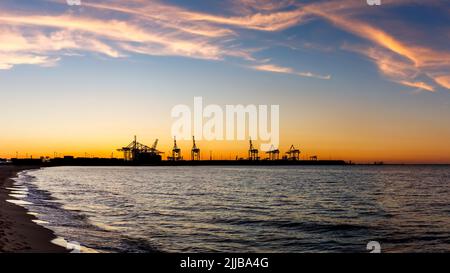 Sonnenuntergang über dem Hafen in Danzig mit Handelsdock und Kränen. Blick vom Stogi Strand in der Ostsee. Silhouette des Industriehafens Stockfoto