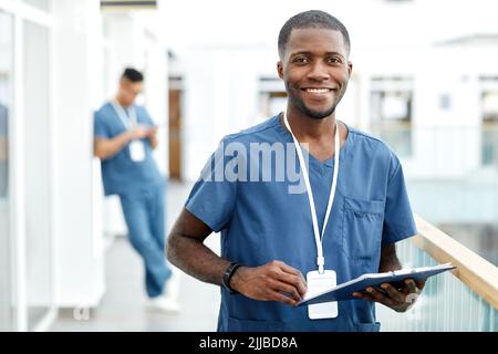 Waist-up-Porträt eines jungen afroamerikanischen Arztes, der mit der Kamera lächelt, während er in der Halle einer modernen Klinik steht und Platz kopiert Stockfoto
