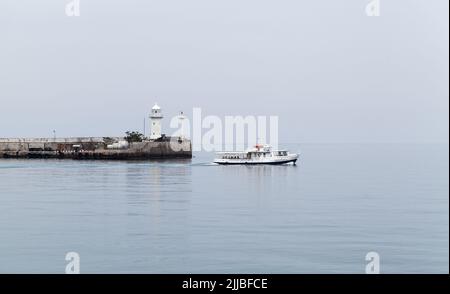 Küstenansicht des Hafens von Jalta. An einem bewölkten Tag segelt ein kleines Passagierschiff in der Nähe des weißen Leuchtturmturms Stockfoto